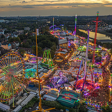 Een foto van de Cranger Kirmes na zonsondergang: je ziet veel verlichte carrousels, kermiswinkels en mensen.