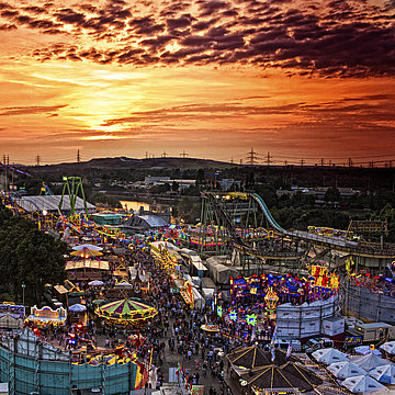 Een foto van de Cranger Kirmes bij zonsondergang: je ziet veel verlichte carrousels, veel mensen en een reuzenrad.