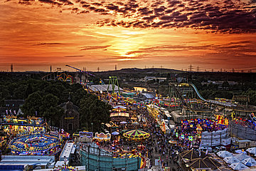 Een foto van de Cranger Kirmes bij zonsondergang: je ziet veel verlichte carrousels, veel mensen en een reuzenrad.